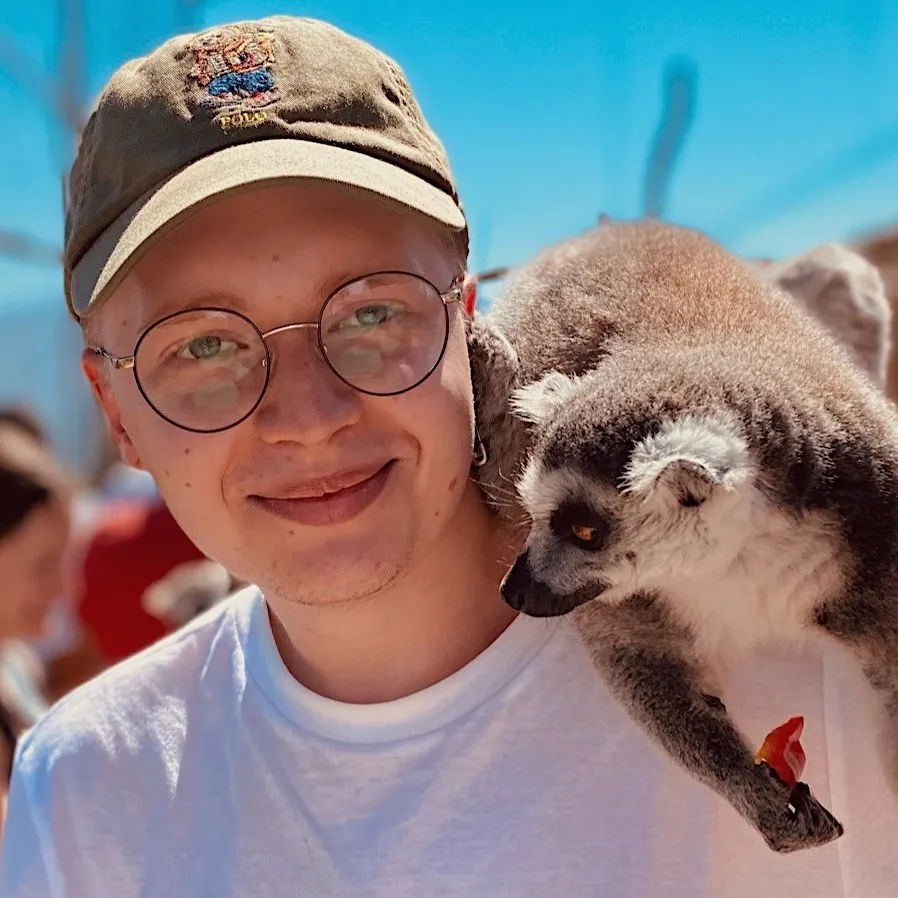 młody mężczyzna w zielonej czapce z daszkiem, białej koszulce, okularach, z lemurem na ramieniu